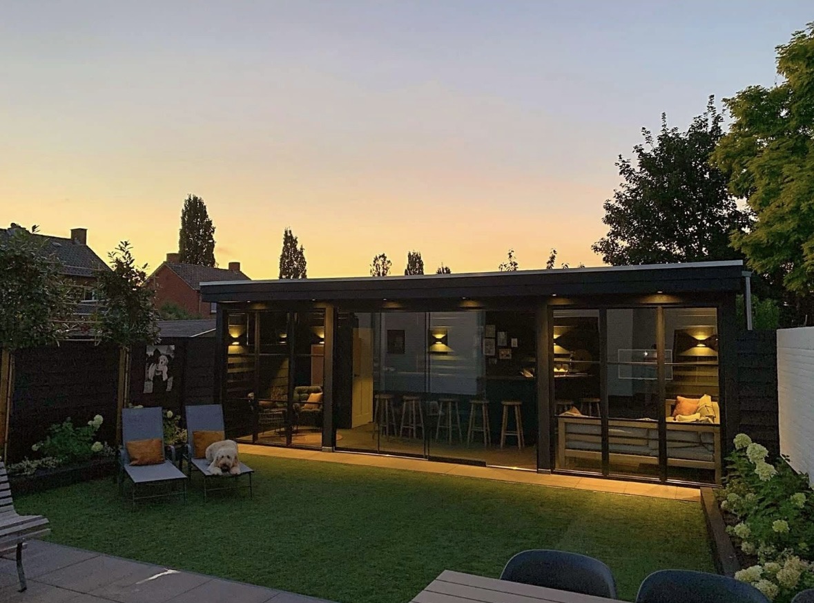 Tuinkamer met glazen schuifwanden en verlichting bij zonsondergang, met gazon en ligstoelen ervoor