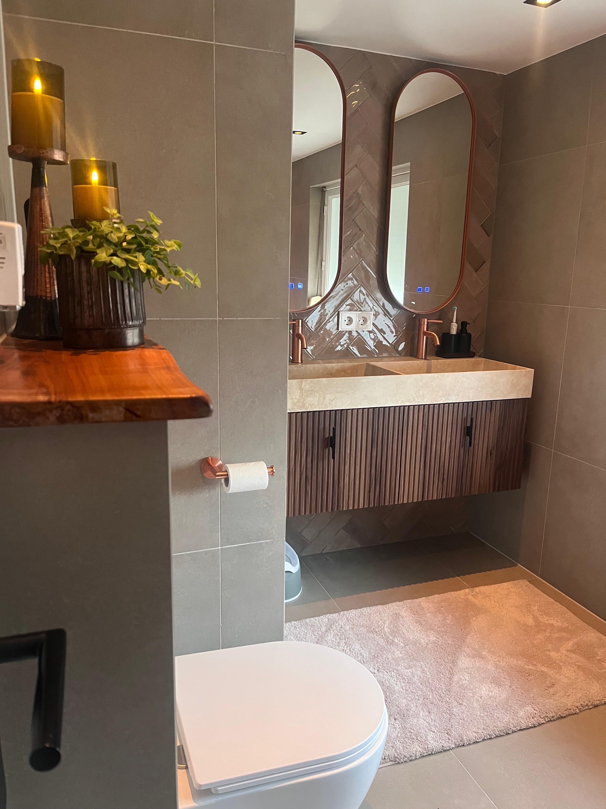 Badkamer met grote grijze tegels, een zwevend houten meubel en twee ovale spiegels