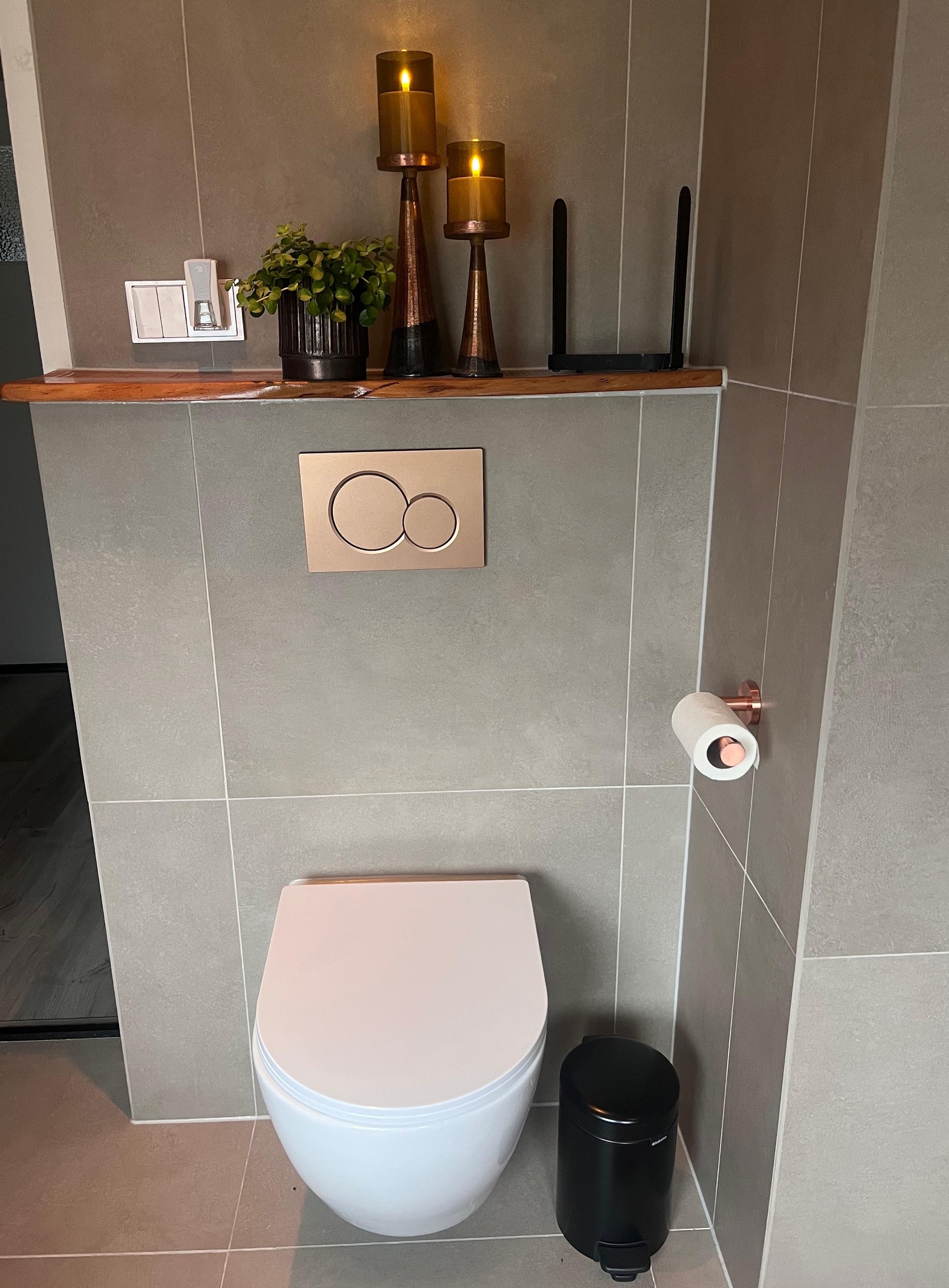 Hangend toilet tegen een betegelde wand met een koperkleurige bedieningsplaat en een houten plank